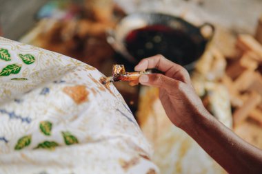 Close up picture of a person's hand drawing the design pattern of Indonesian batik tulis fabric using Canting and malam wax.