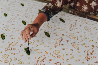 Close up picture of a person's hand drawing the design pattern of Indonesian batik tulis fabric using colorful malam wax.