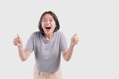 Cute young Asian woman smiling with a funny face and showing a thumb up gesture, isolated by white background.