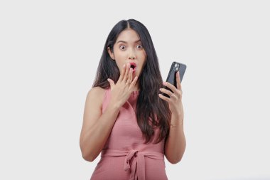 Şok olmuş pembe elbiseli genç bir Asyalı kadın elinde bir akıllı telefon, yüzünde şaşırmış bir ifade var, beyaz arka planla izole edilmiş..