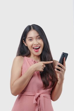 Gülümseyen genç Asyalı kadın elinde tuttuğu akıllı telefonu işaret ediyor. Beyaz arka planla izole edilmiş pembe bir elbise giyiyor..
