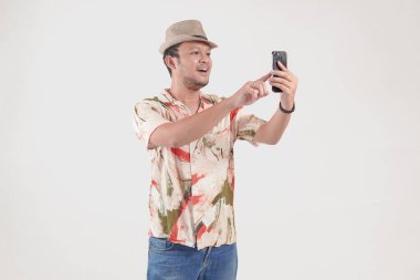 Şapka ve Hawaii tişörtü giyen neşeli Asyalı genç adam, beyaz arka planla izole edilmiş bir gülümsemeyle ona bakarken akıllı telefonunu kullanıyor..