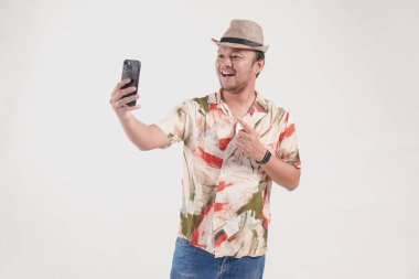 Havai tişörtü giyen neşeli genç Asyalı adam video görüşmesi yapıyor ve telefonu işaret ediyor, beyaz arka planla izole edilmiş..