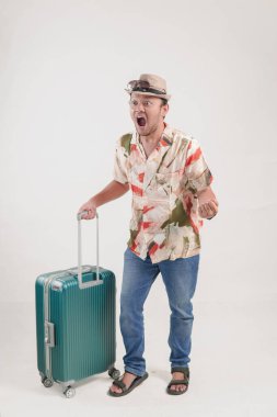 Havai gömlekli heyecanlı genç Asyalı adam izole edilmiş beyaz arka planda bir bavulu tutarken yumruğunu kaldırıyor..