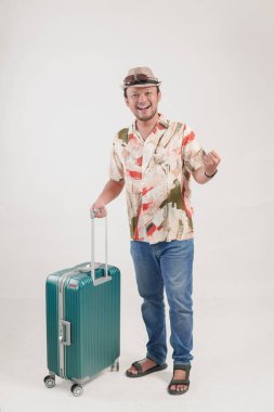 Havai gömlekli başarılı genç Asyalı adam izole edilmiş beyaz arka planda bavulu tutarken elini kaldırıyor..