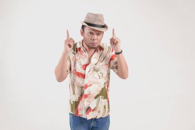 Korkak Asyalı şapkalı ve Hawaii gömlekli genç adam, beyaz arka planla izole edilmiş fotokopi alanını işaret ediyor..