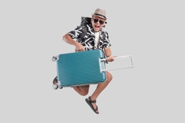 Sırt çantalı ve gözlüklü genç Asyalı turist bavulu izole edilmiş gri arka plandan geçirirken zıplıyor..