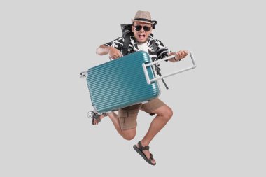 Sırt çantalı ve gözlüklü genç Asyalı turist bavulu izole edilmiş gri arka plandan geçirirken zıplıyor..