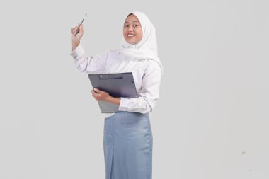 Happy successful Indonesian high school student wearing uniform and hijab holding a clipboard and pen, isolated by white background.