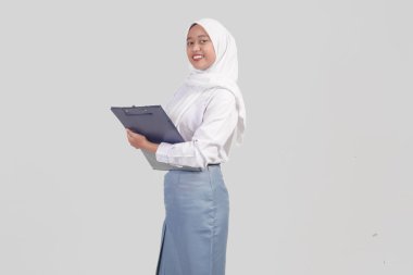 Excited Indonesian high school student wearing uniform and hijab holding a clipboard, isolated by white background.