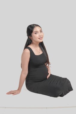 Beautiful pregnant Asian woman is wearing a black dress and sitting on the floor while holding stomach, isolated by white background.