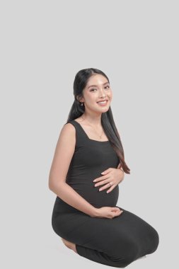 Beautiful pregnant Asian woman sitting on the floor, holding stomach while smiling happily, isolated by white background.