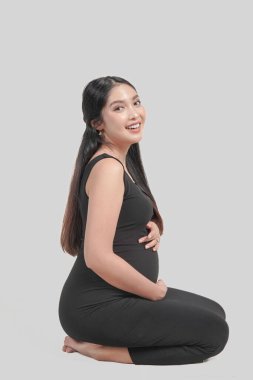 Beautiful pregnant Asian woman sitting on the floor, holding stomach while smiling happily, isolated by white background.