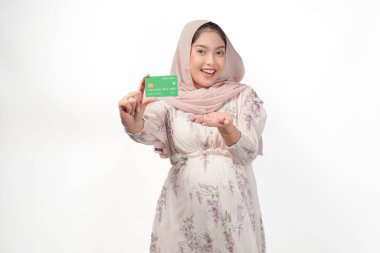 Sırıtkan Asyalı hamile kadın tesettür içinde kredi veya banka kartı sunuyor, beyaz arkaplanla izole edilmiş..