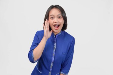 Young Asian customer service woman in blue uniform covering the side of her face to talk secret gossip, isolated by white background.