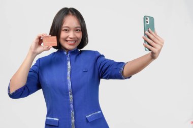 Cheerful Asian customer service woman in blue uniform taking a selfie while holding credit or debit card, isolated by white background.