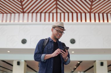 Smiling young Asian man with a backpack is holding smartphone waiting at airport lounge lobby.