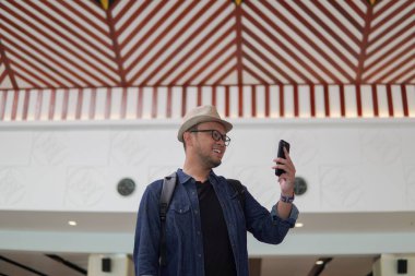 Smiling young Asian man with a backpack is holding smartphone waiting at airport lounge lobby.