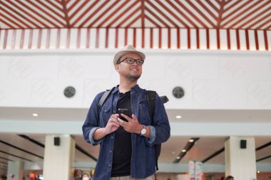 Smiling young Asian man with a backpack is holding smartphone waiting at airport lounge lobby.