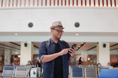 Smiling young Asian man with a backpack is holding smartphone waiting at airport lounge lobby.