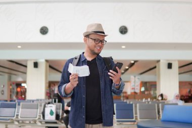 Excited young Asian man bringing backpack smiling while holding a ticket and a smartphone to go on a holiday.