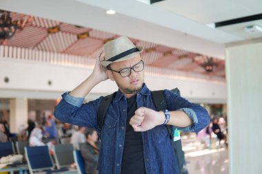 Handsome young Asian man checking time on watch, feeling upset while waiting in an airport lounge because of delayed schedule.