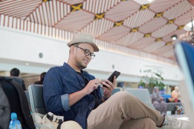 Cheerful young Asian man sitting on a bench while holding smartphone, waiting at airport lounge lobby.