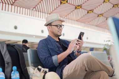 Cheerful young Asian man sitting on a bench while holding smartphone, waiting at airport lounge lobby.