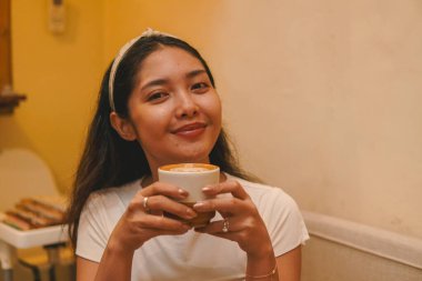 Smiling young Asian woman holding a coffee cup, taking a break and relaxing in a cafe.