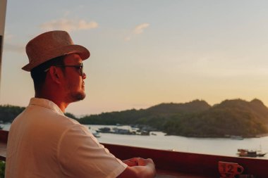 Handsome young Asian man wearing sunglasses smiling and enjoying the sunset scenery at Labuan Bajo beach.