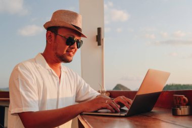 Serious young Asian man working from cafe while enjoying the sunset scenery at Labuan Bajo beach.