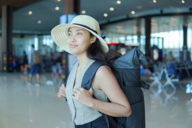 Smiling young Asian woman wearing straw hat and backpack to go on a holiday, waiting at the airport.