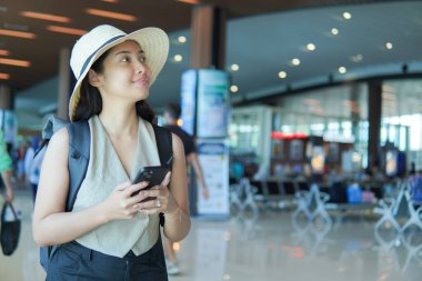 Joyful young Asian woman wearing straw hat and backpack, waiting at the airport while holding her smartphone.