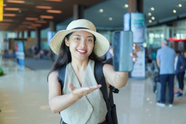 Smiling young Asian woman with straw hat and backpack at airport lobby, pointing at blank screen on her smartphone.