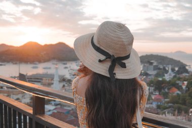 Back view of a young Asian woman wearing a straw hat is looking out over Labuan Bajo city and ocean.