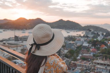 Back view of a young Asian woman wearing a straw hat is looking out over Labuan Bajo city and ocean.