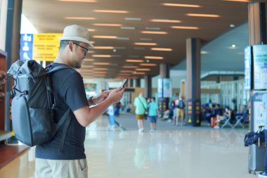 Back view of a young Asian man wearing backpack while looking at his smartphone, waiting at airport lounge lobby.