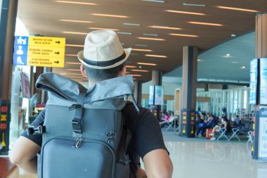 Back view of a young Asian man wearing backpack while looking at his smartphone, waiting at airport lounge lobby.