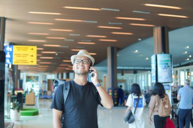 Smiling young Asian man with a backpack using his smartphone to call while waiting at airport lounge lobby.