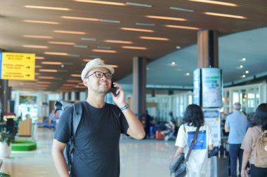 Smiling young Asian man with a backpack using his smartphone to call while waiting at airport lounge lobby.