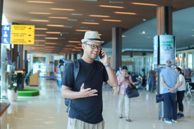Smiling young Asian man with a backpack using his smartphone to call while waiting at airport lounge lobby.