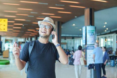 Smiling young Asian man with a backpack using his smartphone to call while waiting at airport lounge lobby.