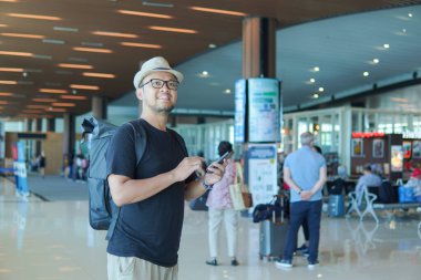 Happy young Asian man with a backpack is holding smartphone waiting at airport lounge lobby.