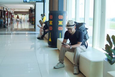 Cheerful young Asian man sitting on a bench while holding smartphone, looking out to the window of the airport lounge lobby.