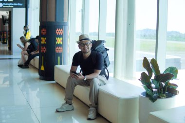 Cheerful young Asian man sitting on a bench while holding smartphone, looking out to the window of the airport lounge lobby.