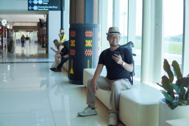 Cheerful young Asian man sitting on a bench while holding smartphone, looking out to the window of the airport lounge lobby.