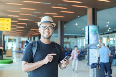 Joyful young Asian man with a backpack is smiling while holding smartphone, waiting at airport lounge lobby.