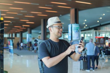 Handsome young Asian man with a backpack is using his smartphone while waiting at airport lounge lobby.