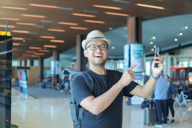 Excited young Asian man with a backpack is using and pointing to his smartphone while waiting at the airport lounge lobby.