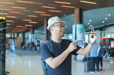Excited young Asian man with a backpack is using and pointing to his smartphone while waiting at the airport lounge lobby.